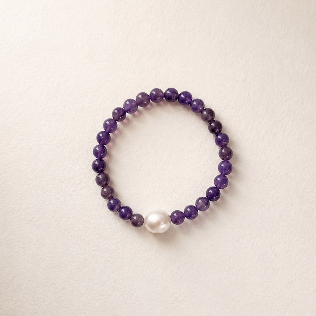 A beaded bracelet featuring dark purple amethyst beads and a single south sea white pearl accent, displayed on a light beige textured background.