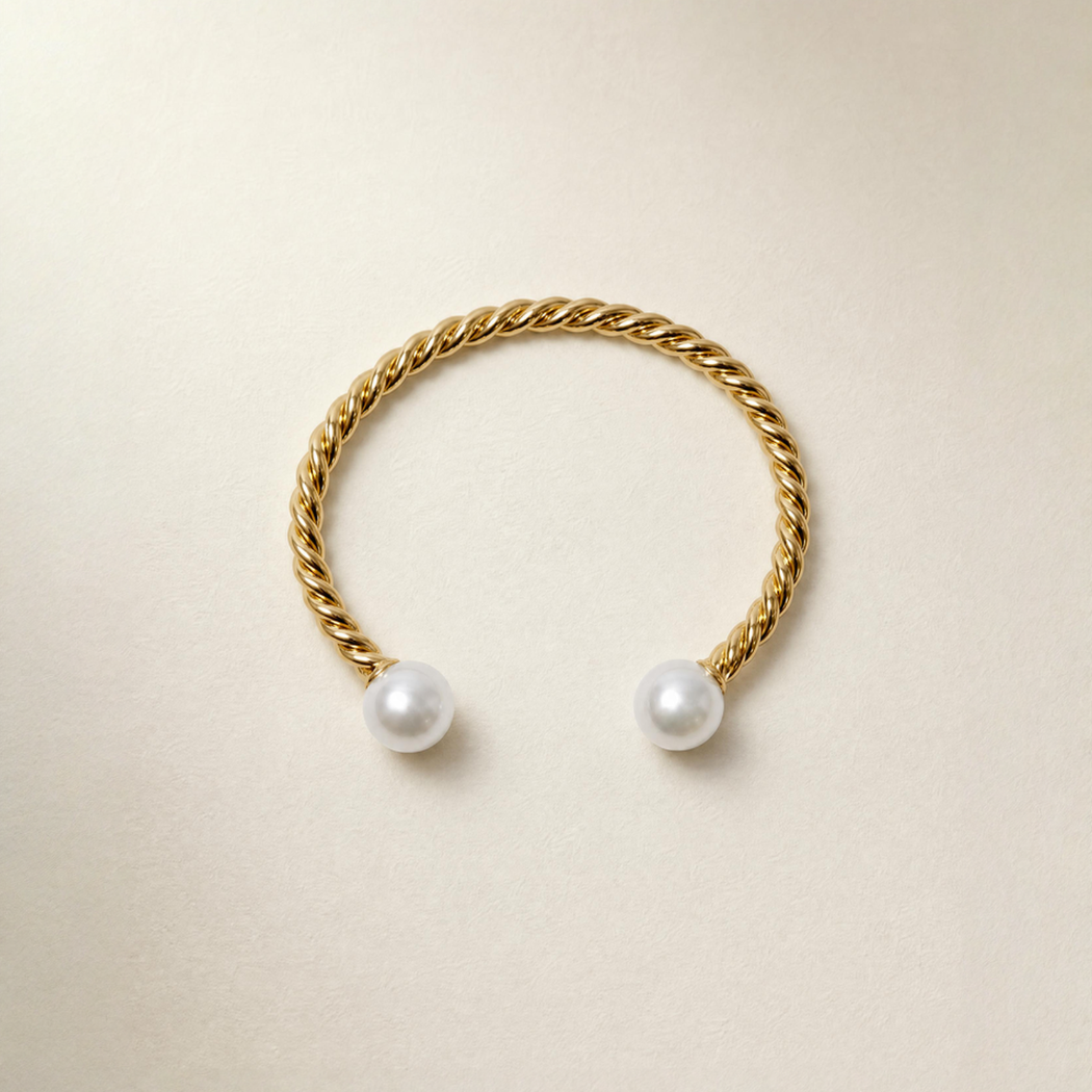 A gold twisted rope cuff ring with two white south sea pearl beads at the open ends, displayed on a light beige textured background.