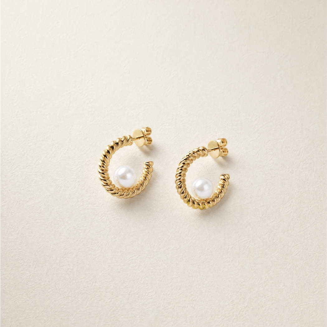 A pair of gold huggie hoop earrings with a twisted rope texture and a small south sea white pearl set in the center, displayed on a textured beige background.