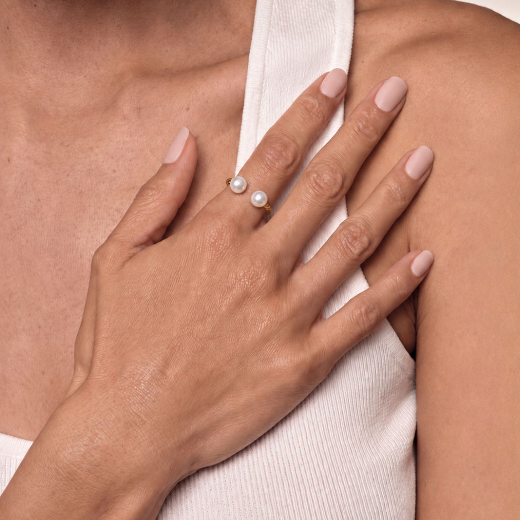 A close-up of a person's hand resting on their shoulder, wearing a delicate gold ring with two south sea white pearls.