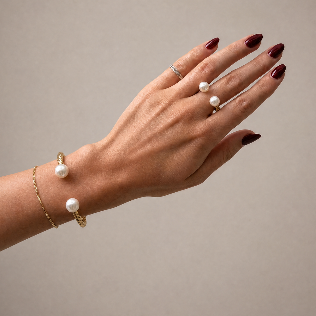 A hand with dark red nail polish wearing a gold cuff bracelet with two white south sea pearls and matching pearl rings on a neutral beige background.