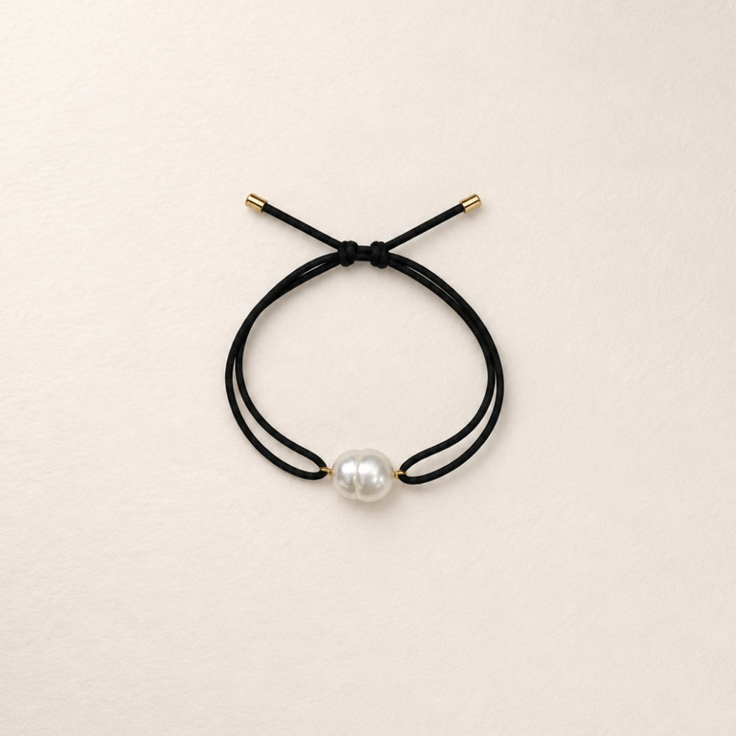 A double-strand black cord bracelet with a single south sea white pearl bead and small gold-toned metal caps, displayed on a light beige textured background.