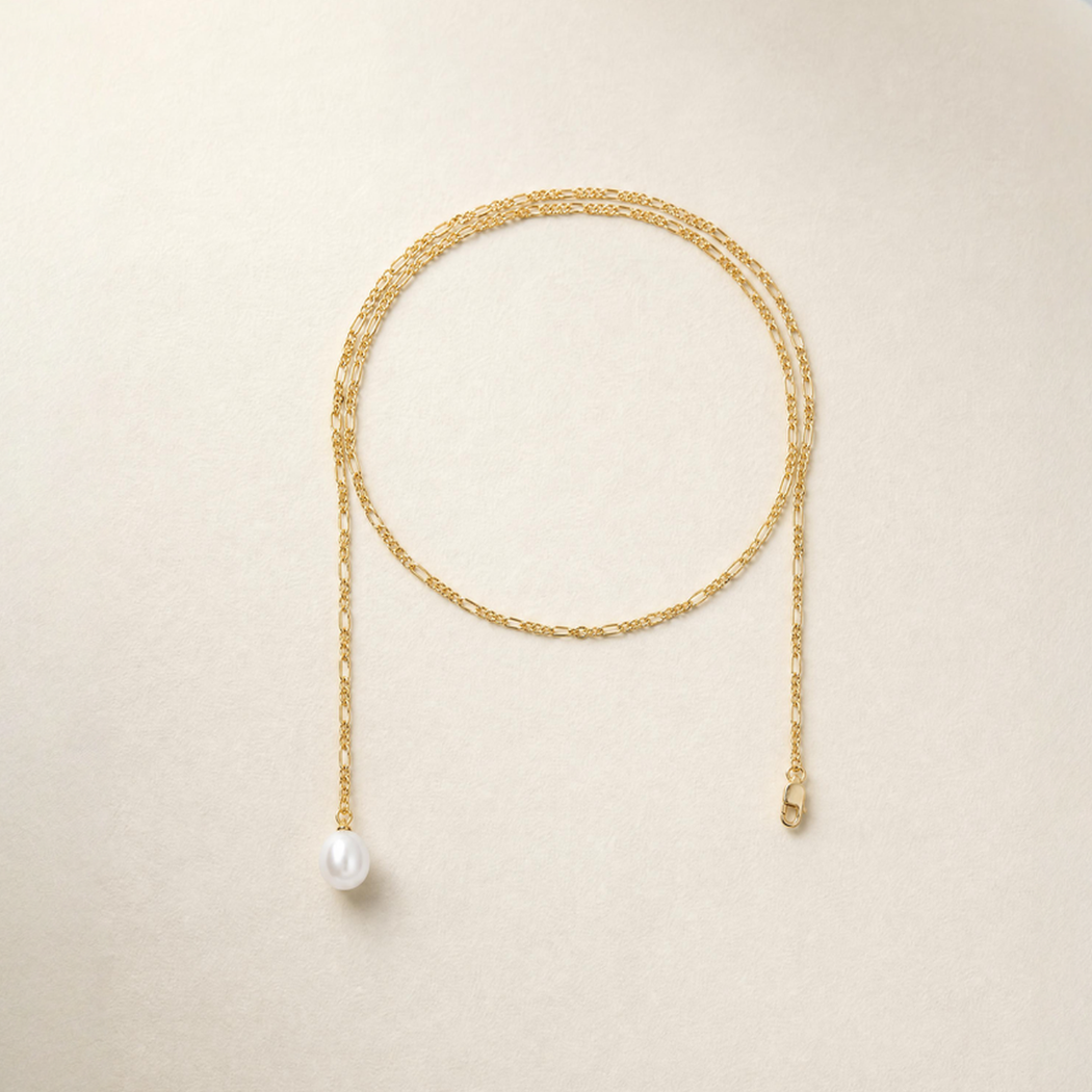 A delicate gold chain anklet with a single white pearl pendant, displayed on a light beige textured background.