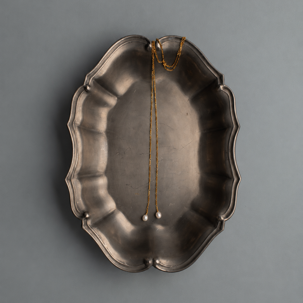 A vintage silver-toned metal platter with a scalloped edge, shown from a top-down perspective on a solid gray background. A gold chain with pearl-like beads is draped over the center of the platter.
