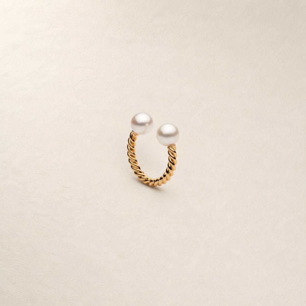 A gold twisted rope ring with two white south sea pearls, displayed on a textured off-white background.