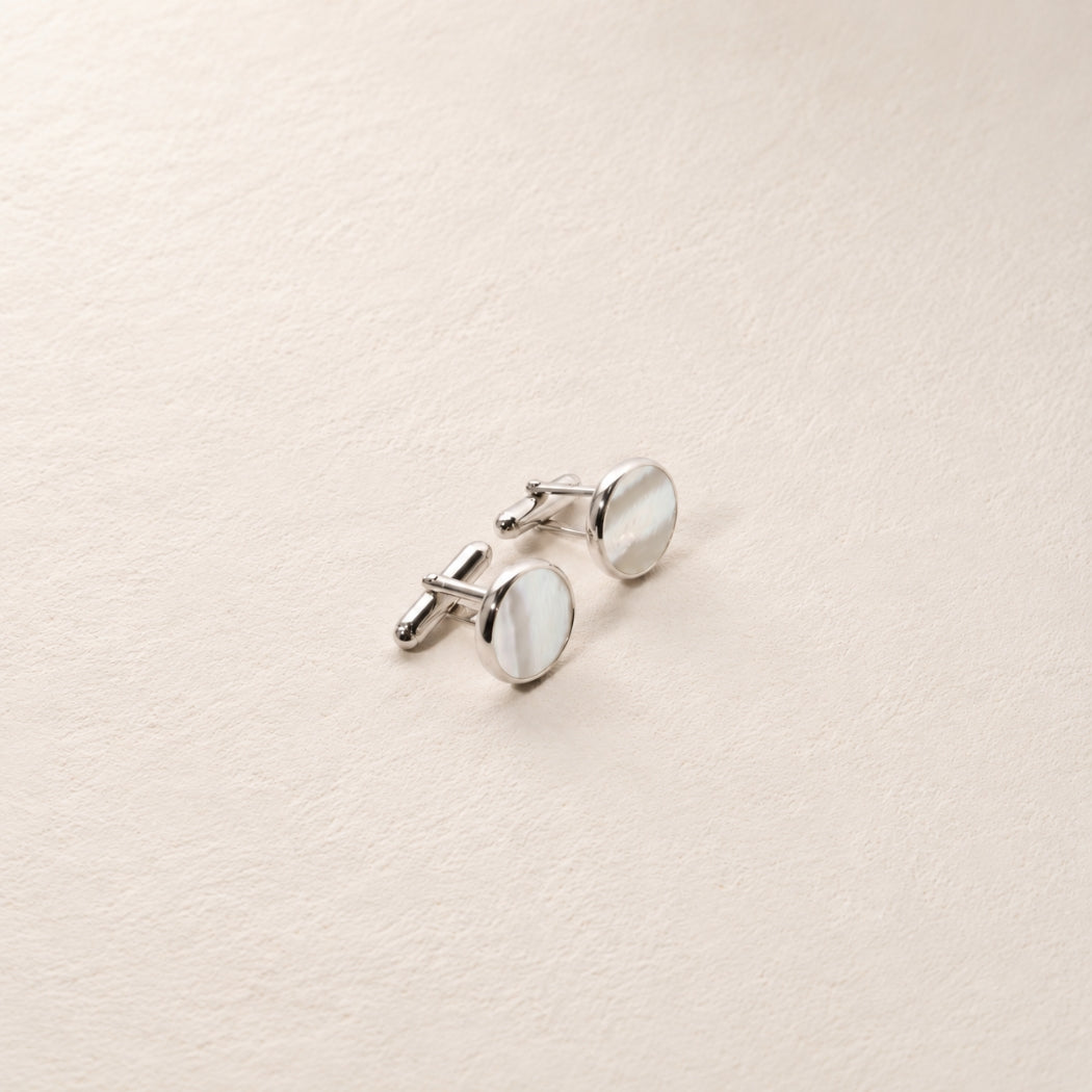 A pair of silver-toned oval cufflinks with white mother-of-pearl inlay, displayed on a textured beige surface.
