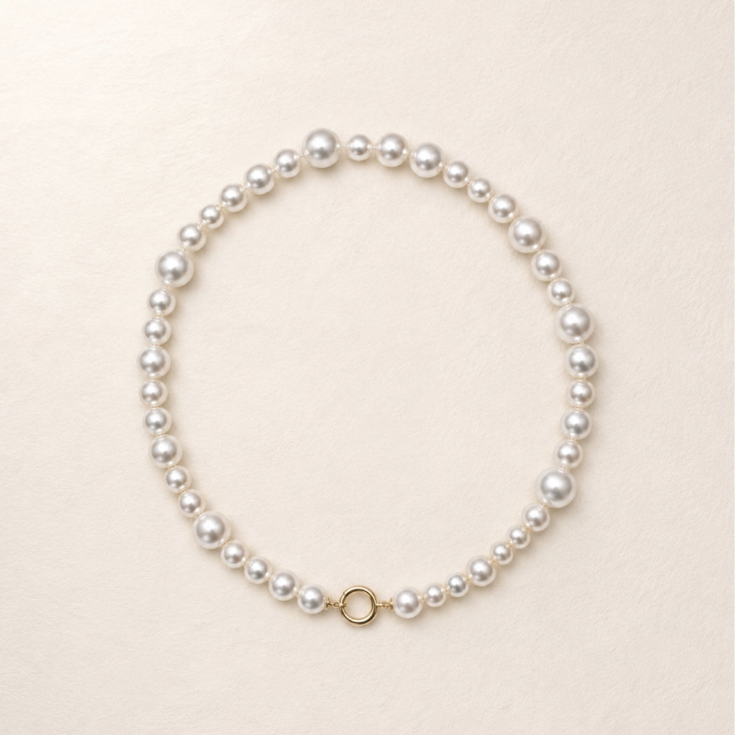 A single-strand south sea pearl necklace with a gold clasp, arranged in a neat circle on a light beige textured background.