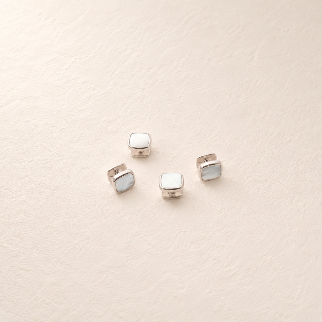 Four silver shirt studs with square mother of pearl inlay, arranged on a textured beige surface.