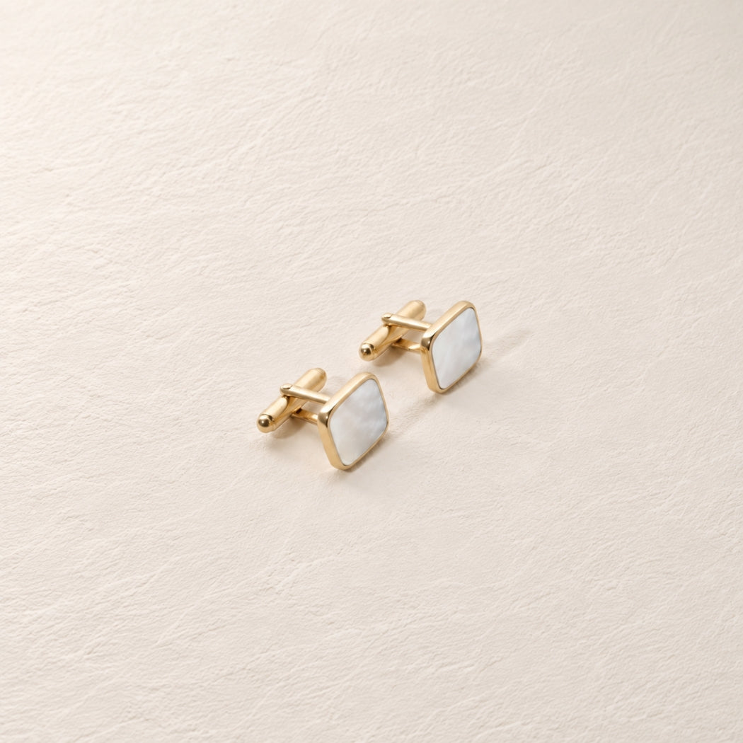 A pair of gold square cufflinks with white mother-of-pearl inlays, displayed on a textured beige surface.