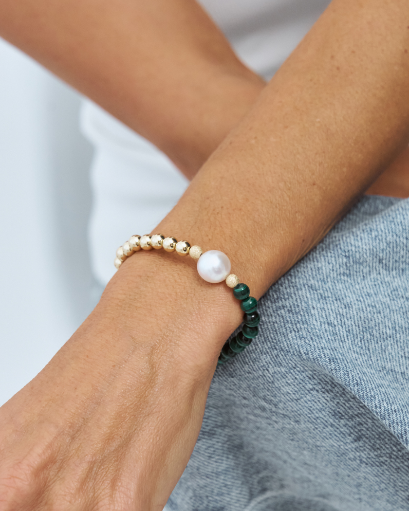 Close-up of a wrist, wearing a bracelet with a south sea pearl, malachite and gold beads on a neutral background