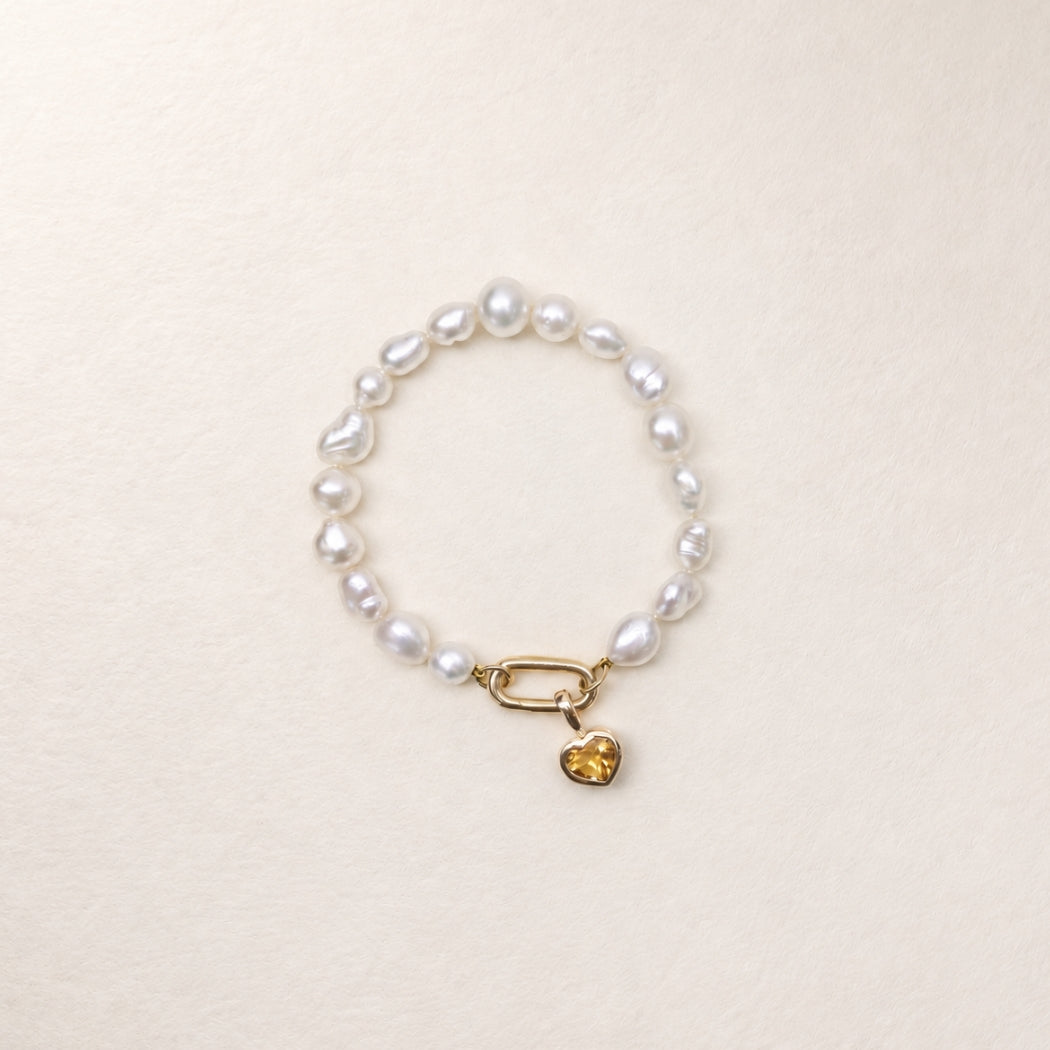 A south sea keshi pearl bracelet, featuring a citrine gold heart charm, displayed on a textured cream background.