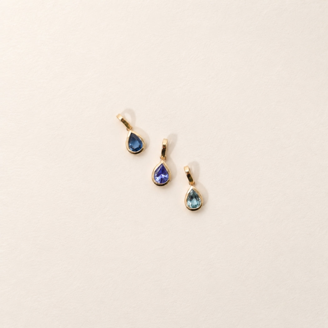 Three gold teardrop pendant charms, blue sapphire, tanzanite and aquamarine. displayed on a light beige textured background.