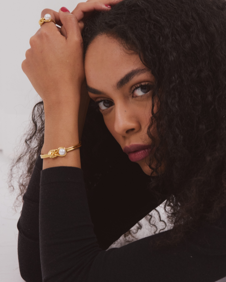 Woman with curly hair wearing a gold bracelet and ring against a white background