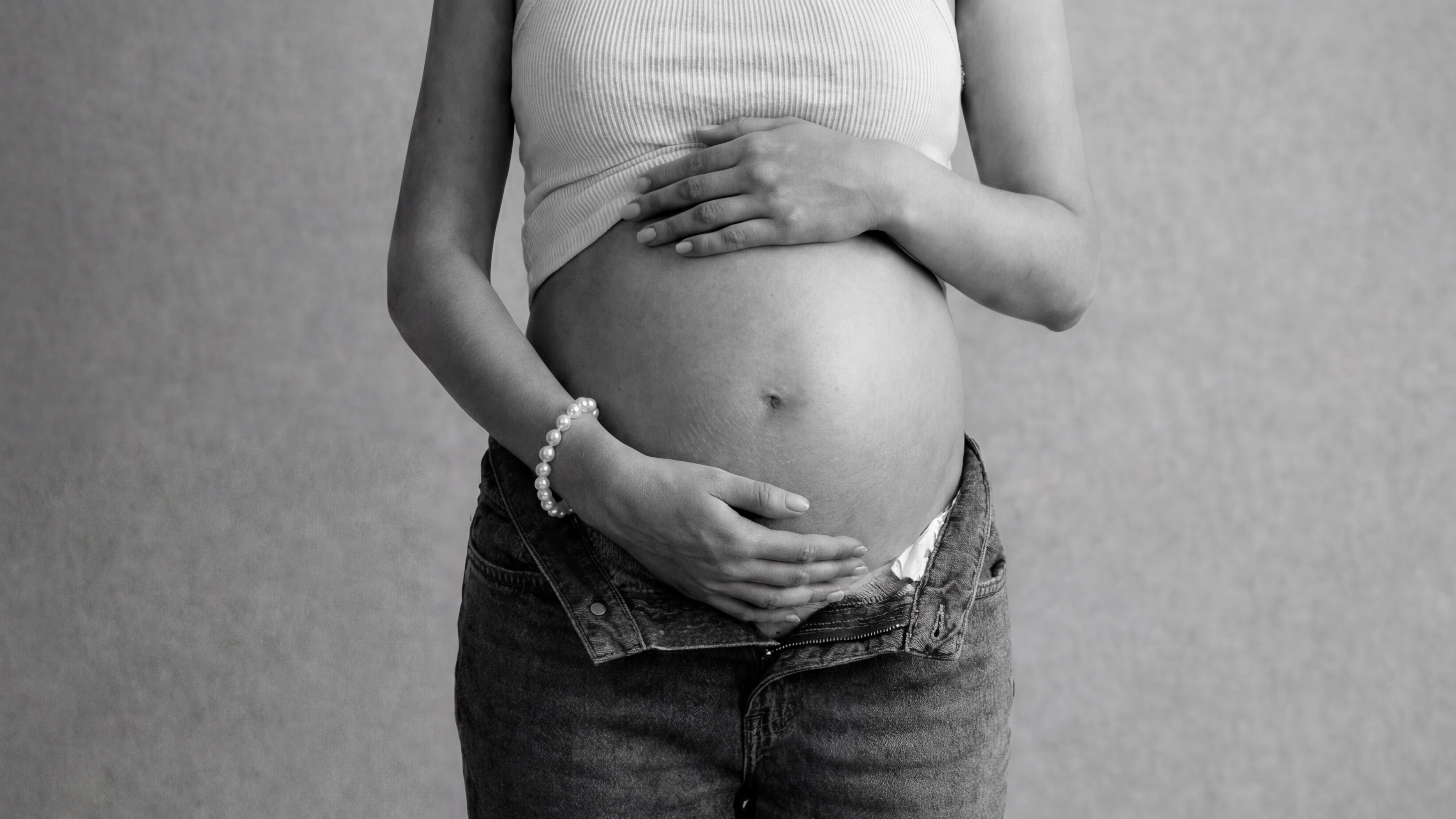Pregnant person wearing a light top and dark jeans against a plain background