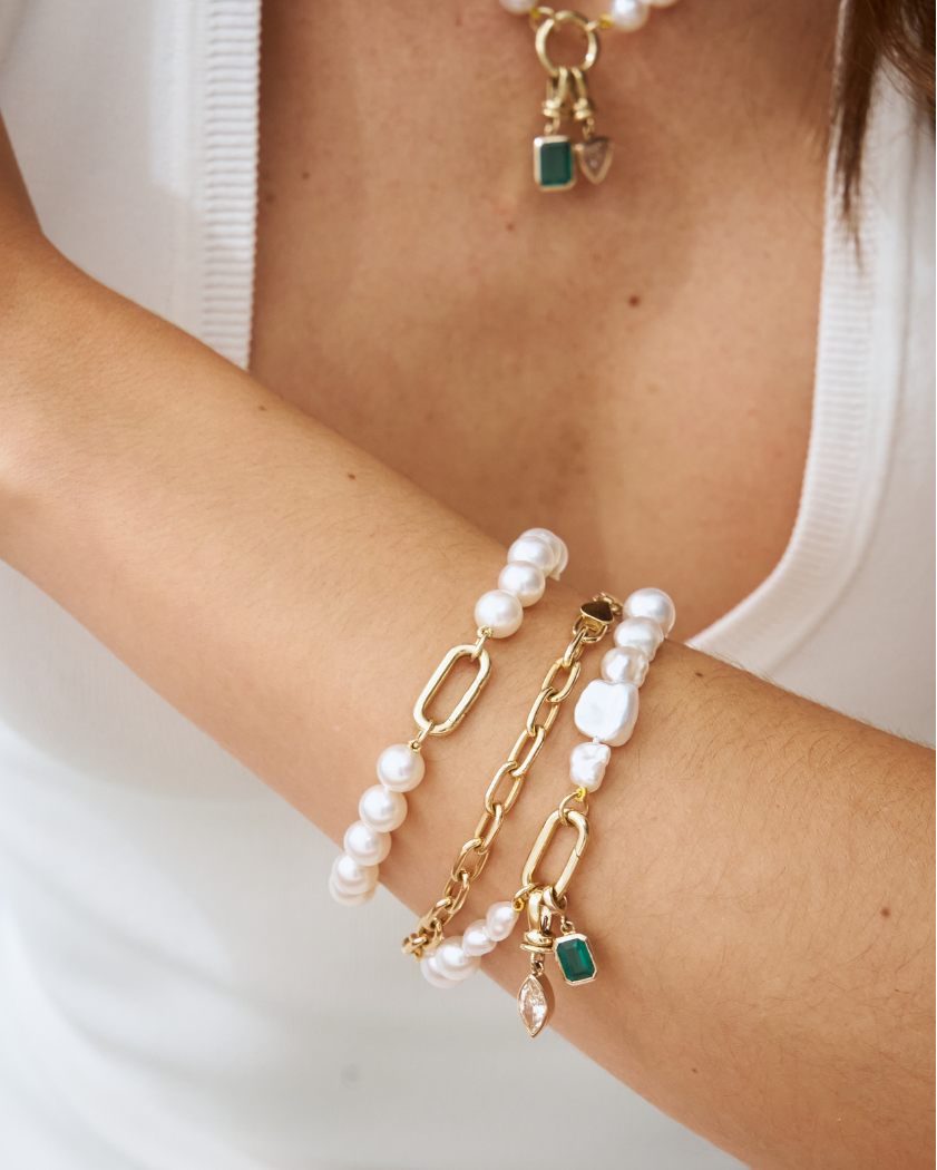 Close-up of a models arm wearing a south sea round pearl bracelet, stacked with a paperclip chain bracelet with emerald and diamond charms.