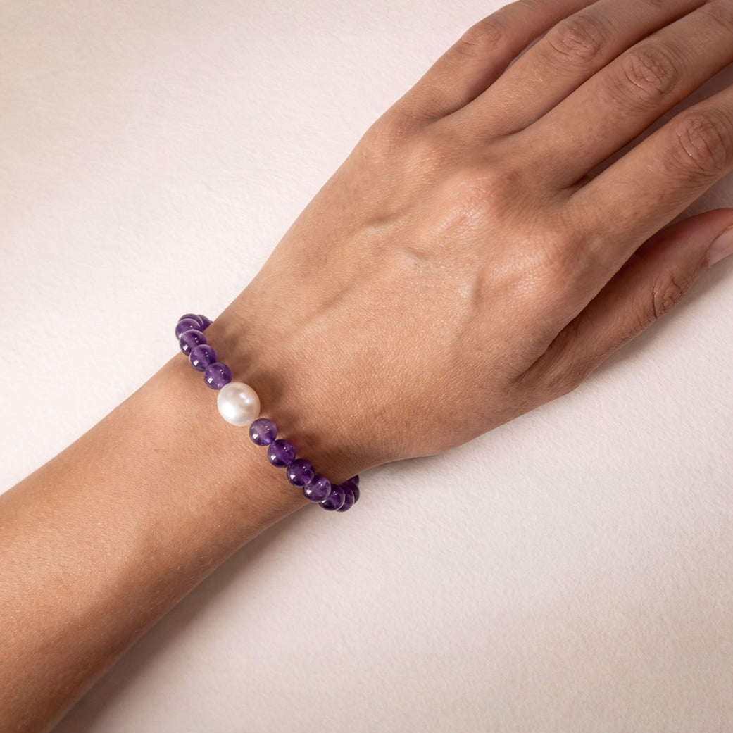 A close-up of a person's wrist wearing a beaded bracelet made of purple amethyst beads with a single south sea white pearl accent.