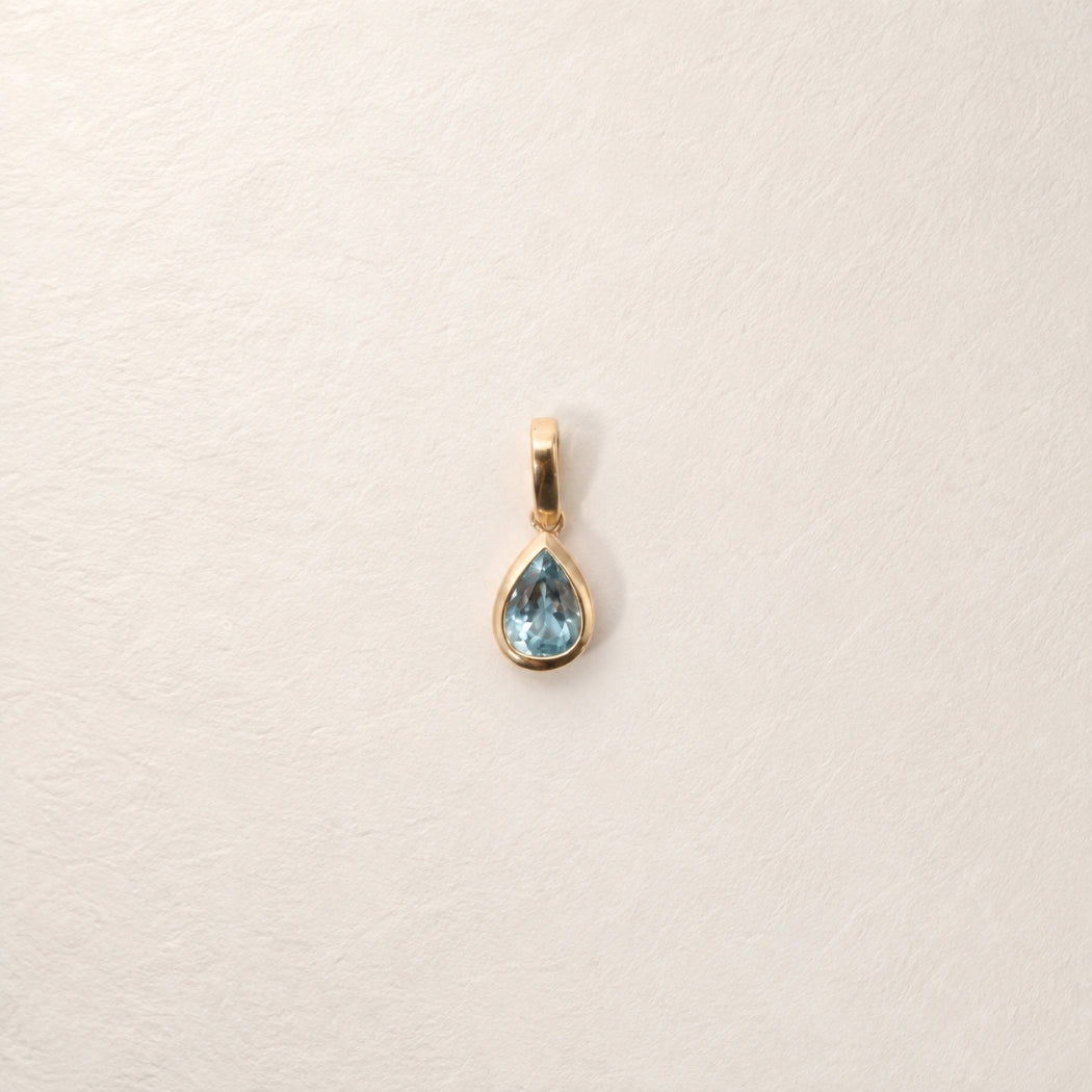A gold pendant with a pear-shaped aquamarine gemstone, displayed on a textured off-white background.