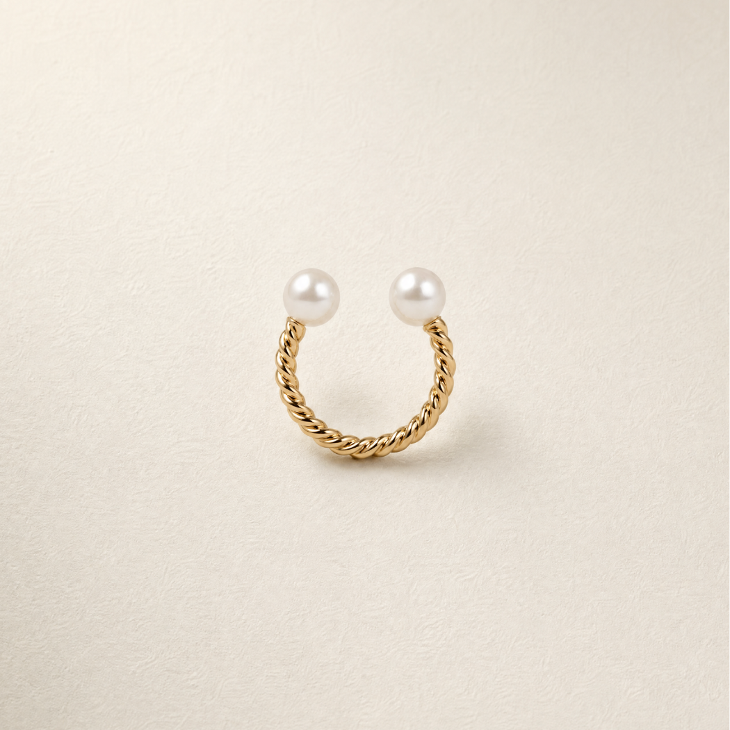 A gold twisted rope ring with two white south sea pearls, displayed on a textured off-white background.