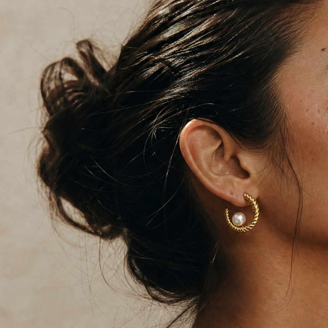 A close-up of a person's ear wearing a gold huggie hoop earring with a south sea white pearl at the center, set against a neutral beige background.