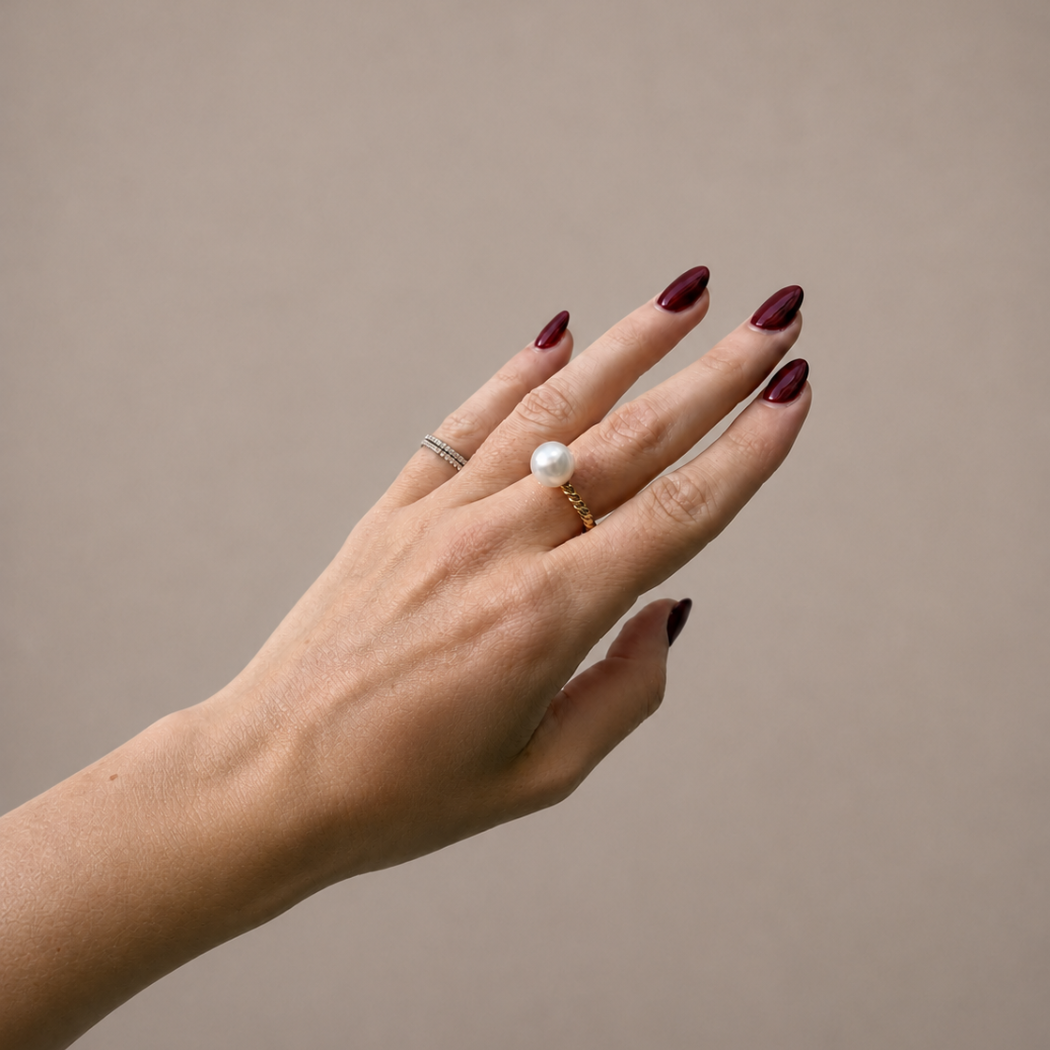 A close-up of a hand with dark burgundy nail polish, showcasing two rings: a delicate silver band on the middle finger and a gold ring with a single white pearl on the ring finger, set against a solid beige background.