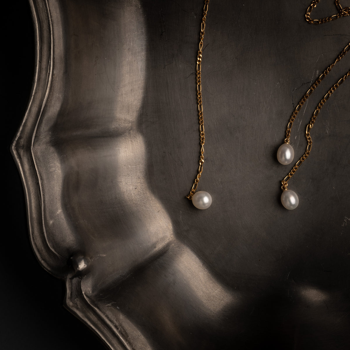 Gold chain necklaces with white pearl pendants displayed on a dark metallic tray against a black background.