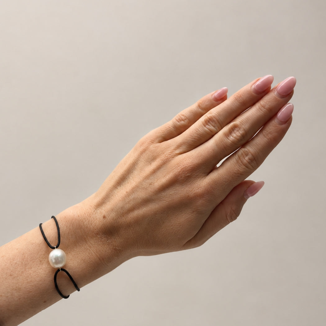 A close-up of a person's hand and wrist wearing a black cord bracelet with a single white south sea pearl bead, set against a plain light gray background.