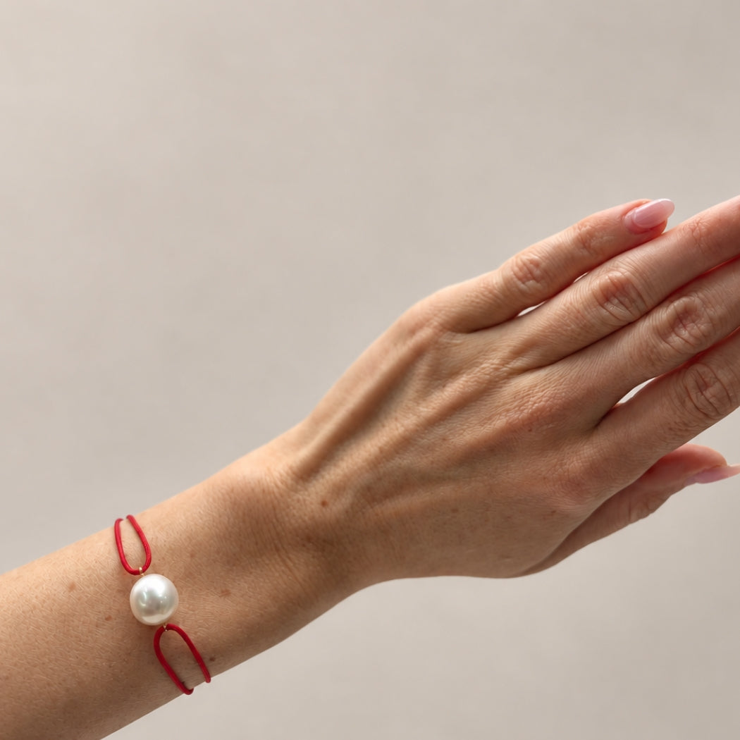 A close-up of a person's wrist wearing a red cord bracelet with a single white south sea pearl bead, set against a plain light gray background.