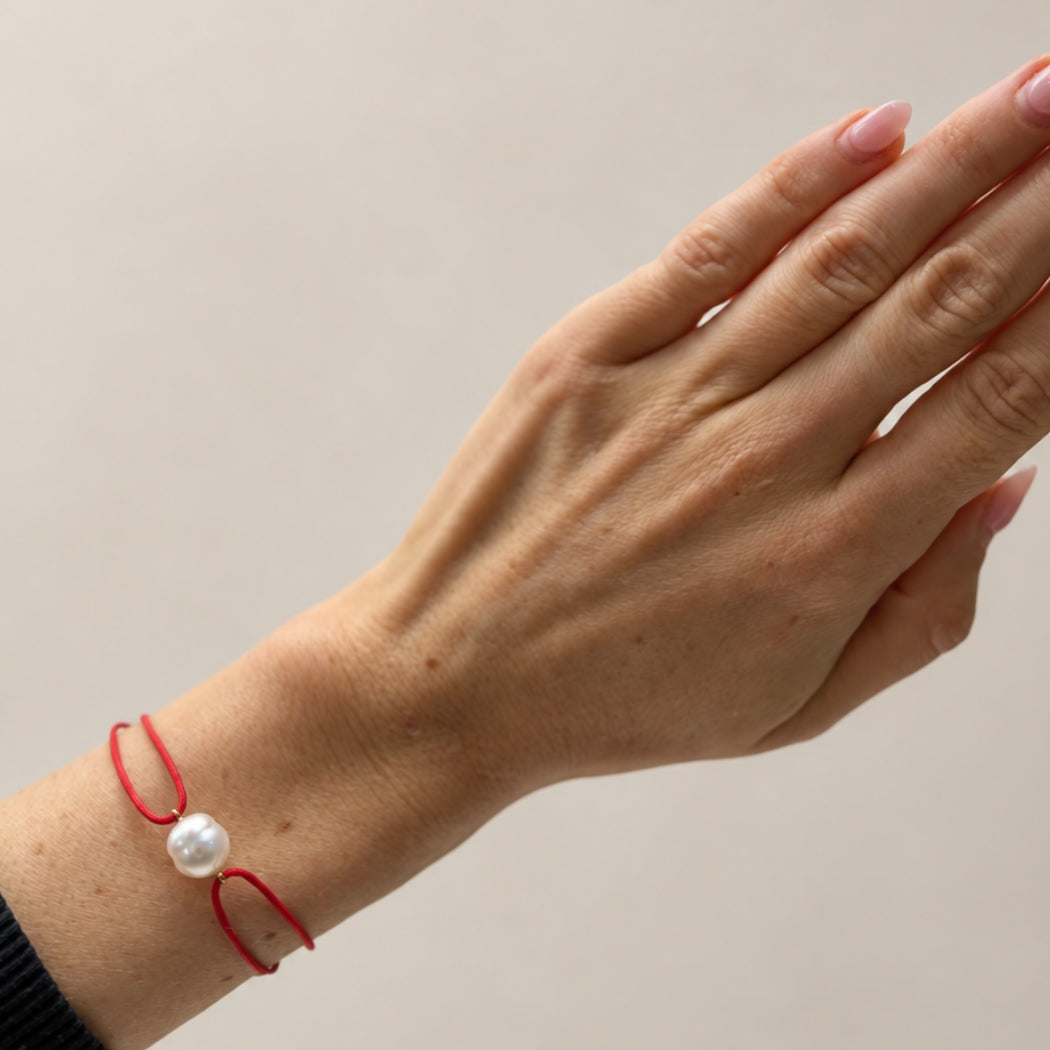 A close-up of a person's hand and wrist wearing a delicate red cord bracelet with a single white south sea pearl.
