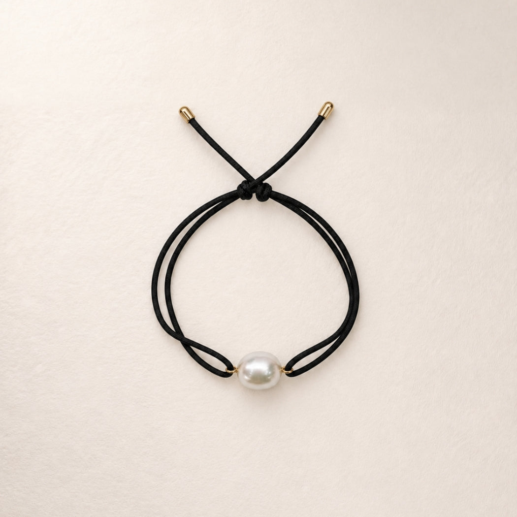 A double-strand black cord bracelet with a single south sea white pearl bead and small gold-toned metal caps, displayed on a light beige textured background.