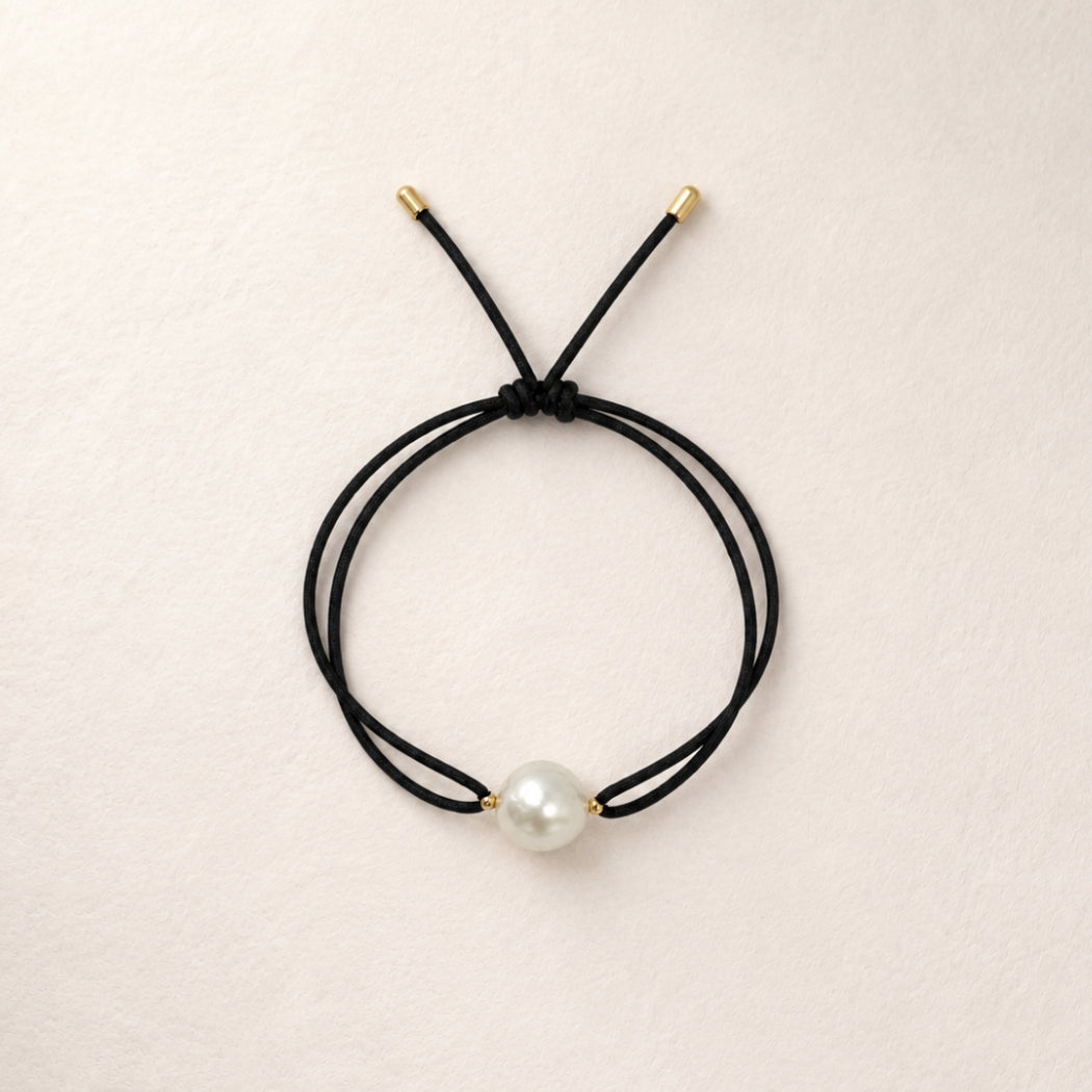 A double-strand black cord bracelet with a single south sea white pearl bead and small gold-toned metal caps, displayed on a light beige textured background.