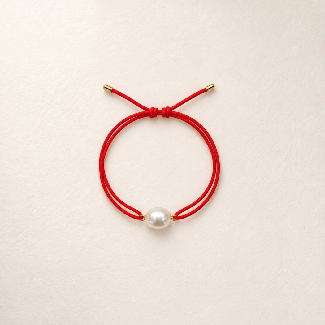A double-strand red cord bracelet with a single south sea white pearl bead and small gold-toned metal caps, displayed on a light beige textured background.