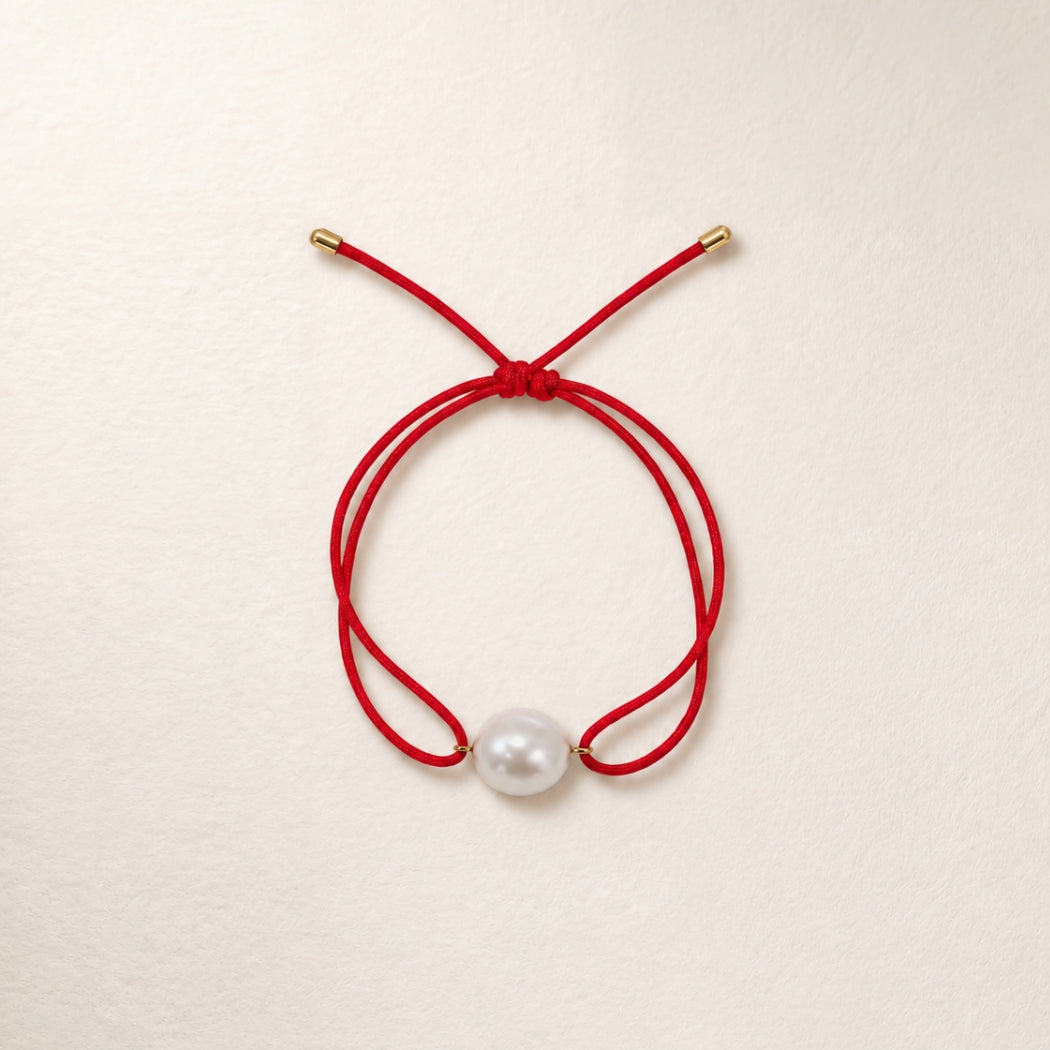A double-strand red cord bracelet with a single south sea white pearl bead and small gold-toned metal caps, displayed on a light beige textured background.