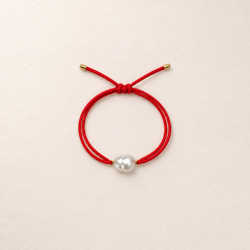 A double-strand red cord bracelet with a single south sea white pearl bead and small gold-toned metal caps, displayed on a light beige textured background.