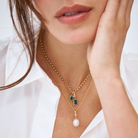 Close-up of a model wearing a 9k gold belcher chain necklace with a small and large emerald charm, layered with a south sea pearl charm. 