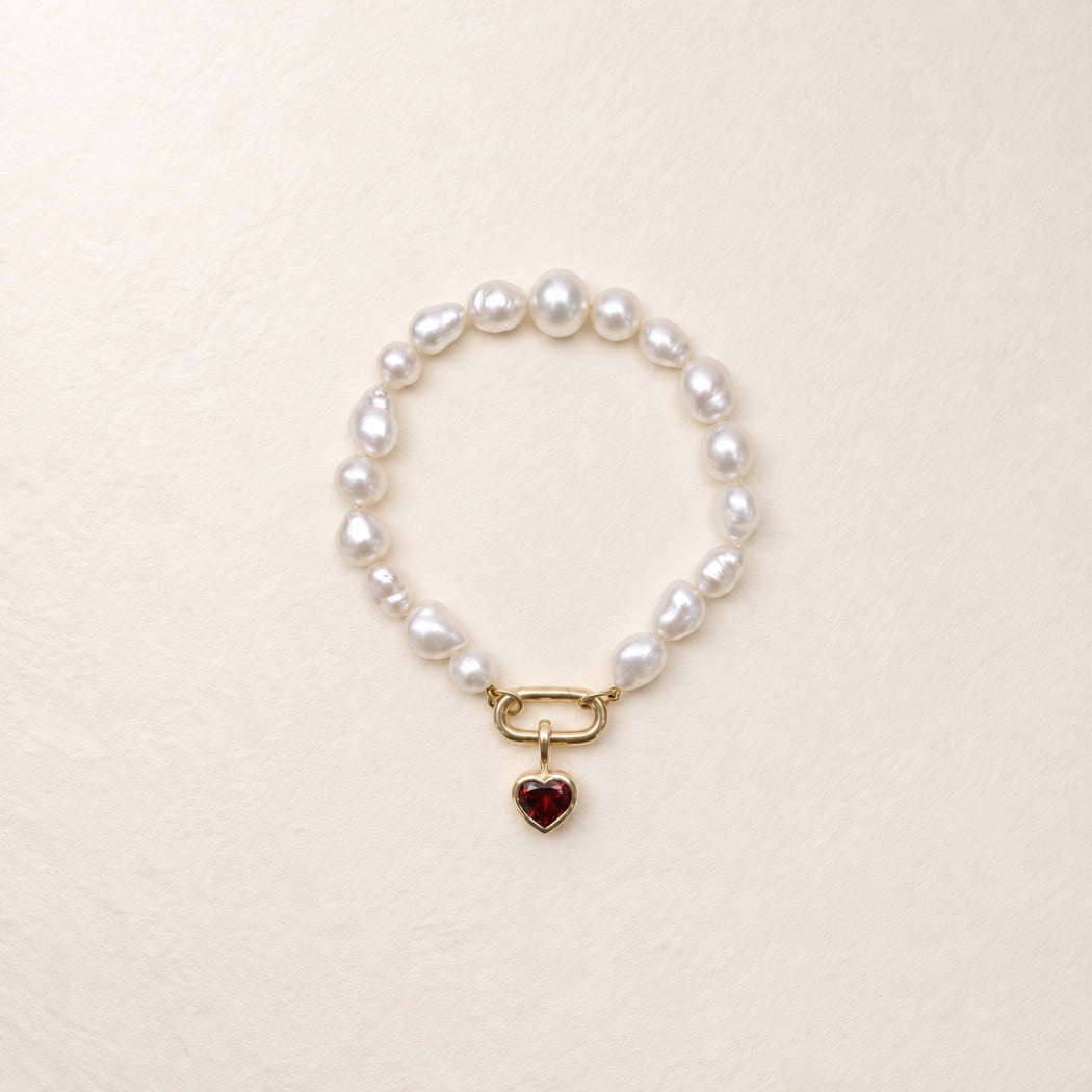 A south sea keshi pearl bracelet with a gold clasp and a dangling red garnet heart charm, displayed on a light beige textured background.