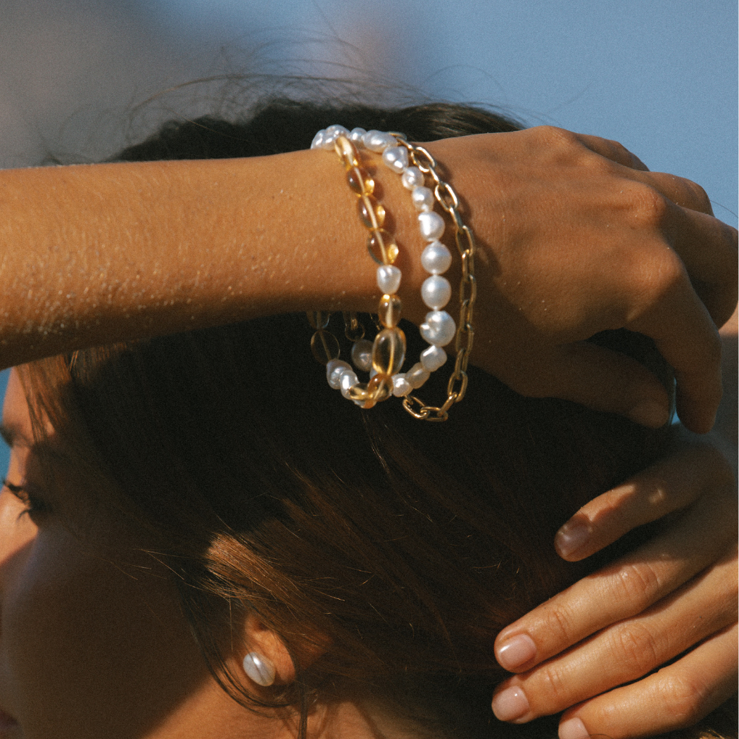 Close-up on arm wearing multiple 9k solid gold South Sea keshi pearl and gemstone bracelets with diamond cushion cut charm.