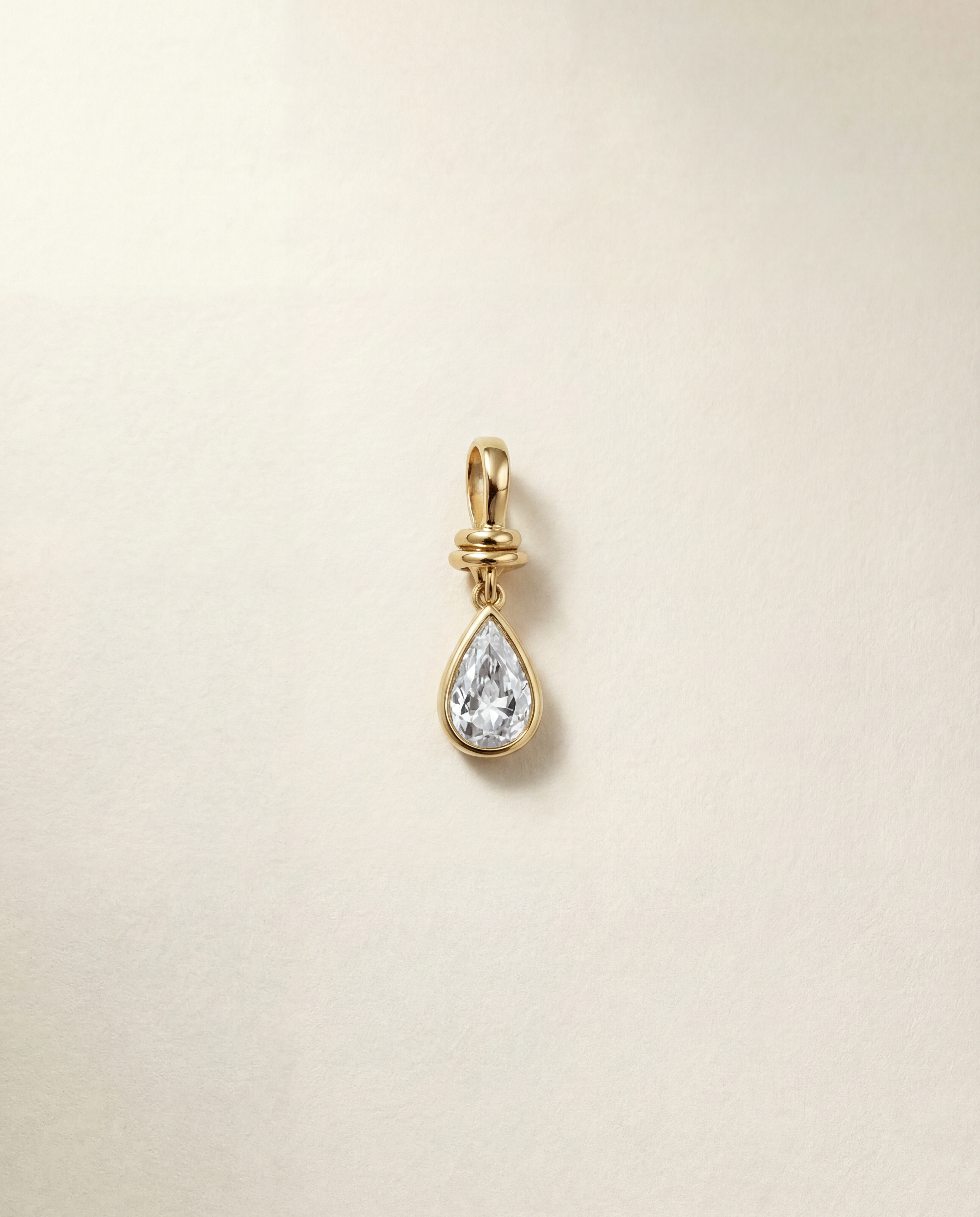 Gold teardrop-shaped pendant with a clear gemstone on a beige background