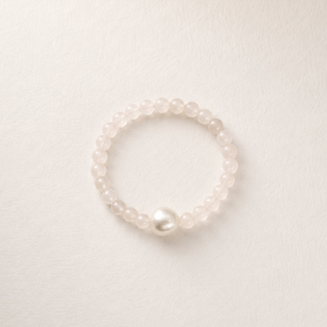 A delicate beaded stretch bracelet featuring rose quartz beads and a single prominent white south sea pearl, displayed on a textured off-white background.
