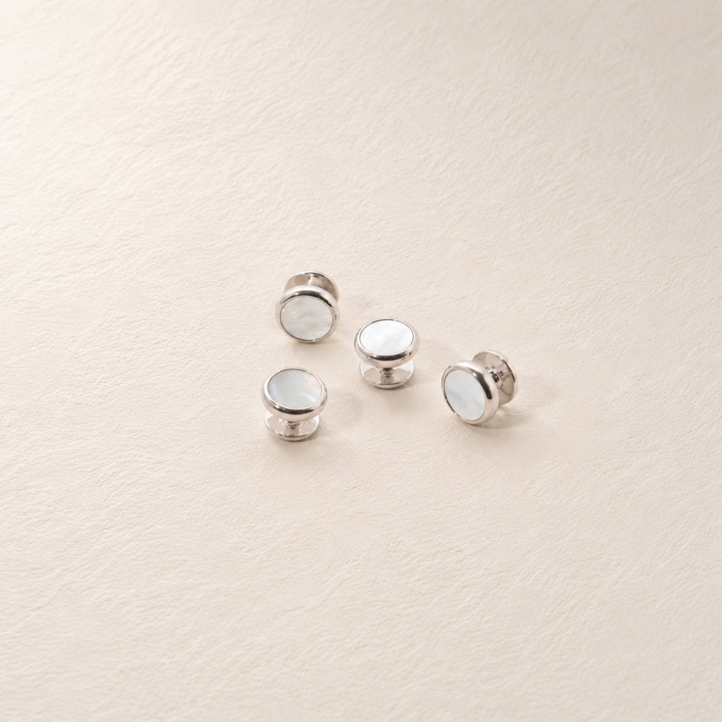 Four silver shirt studs with mother of pearl inlay, arranged on a textured cream background.