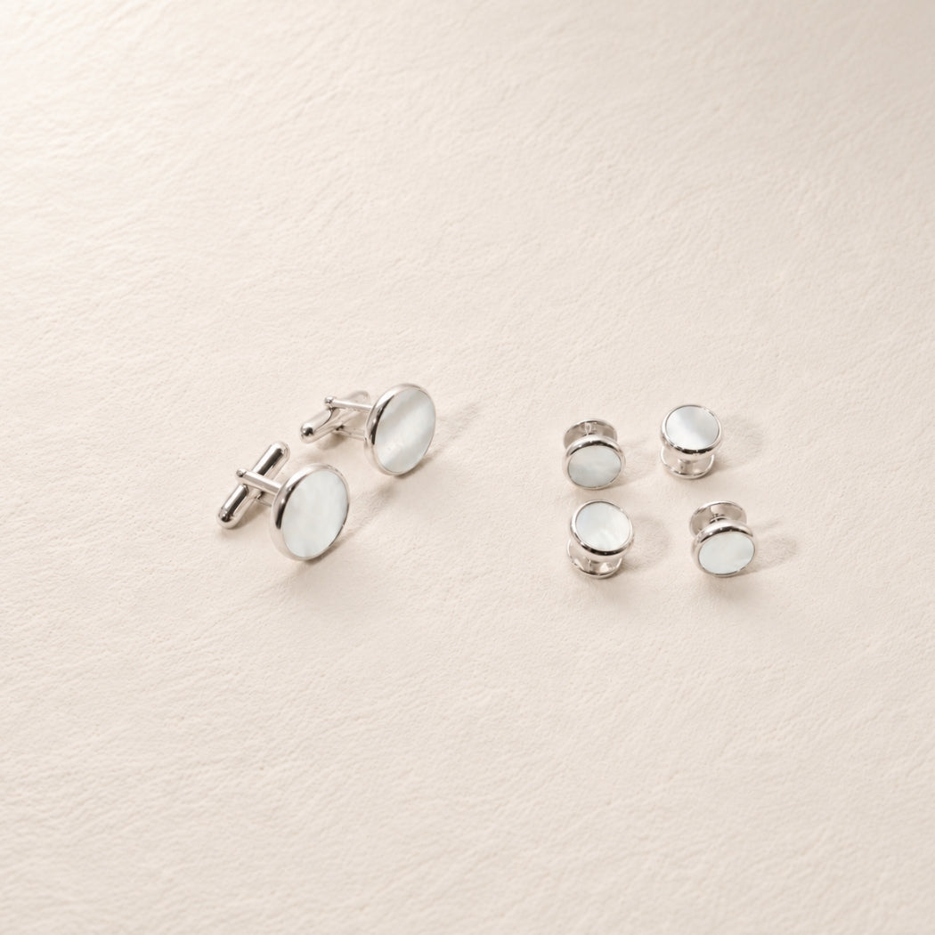 A set of silver cufflinks and matching shirt studs with round white mother of pearl , arranged on a textured off-white background.