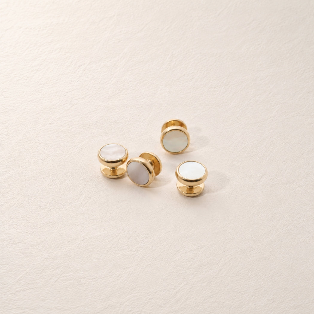 Four small gold-tone shirt studs with mother of pearl inlay, arranged on a light cream textured surface.