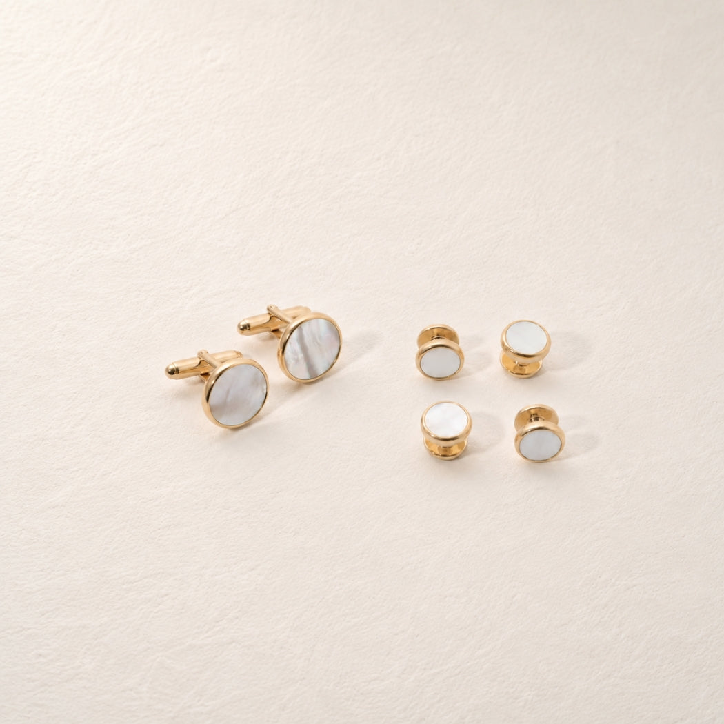 A set of gold-toned cufflinks and matching shirt studs with round white mother of pearl , arranged on a textured off-white background.
