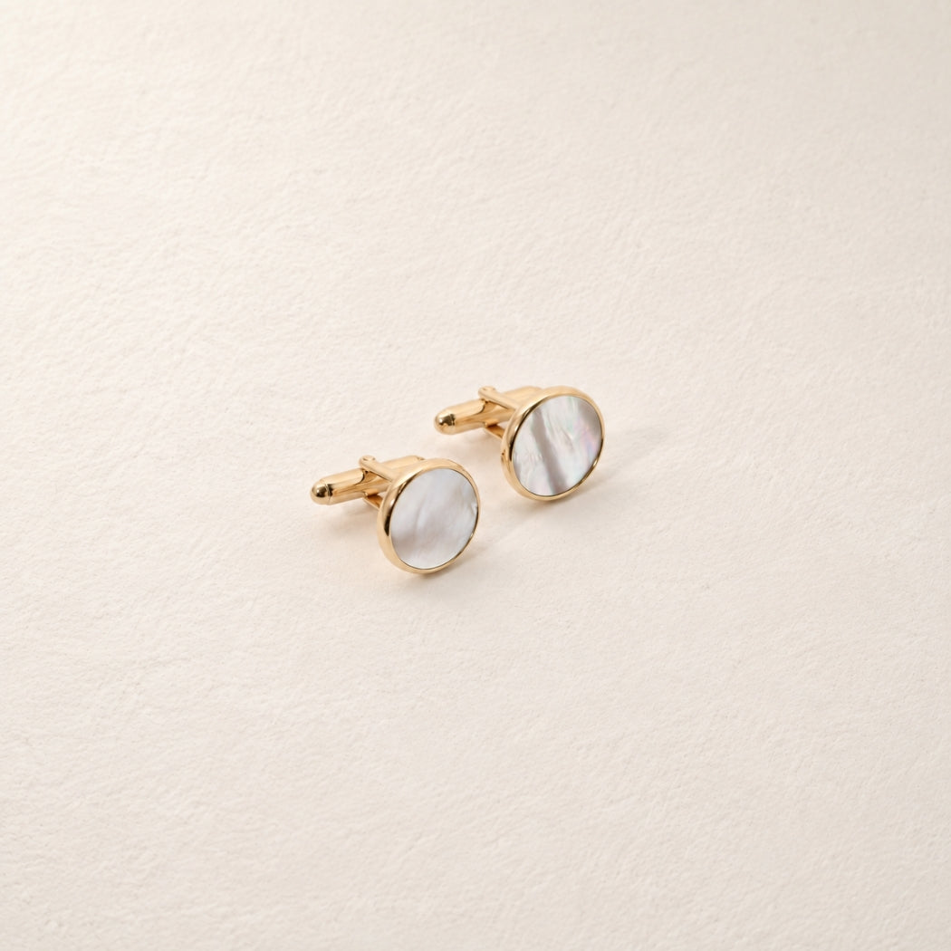 A pair of gold round cufflinks with white mother-of-pearl inlay, displayed on a textured off-white background.