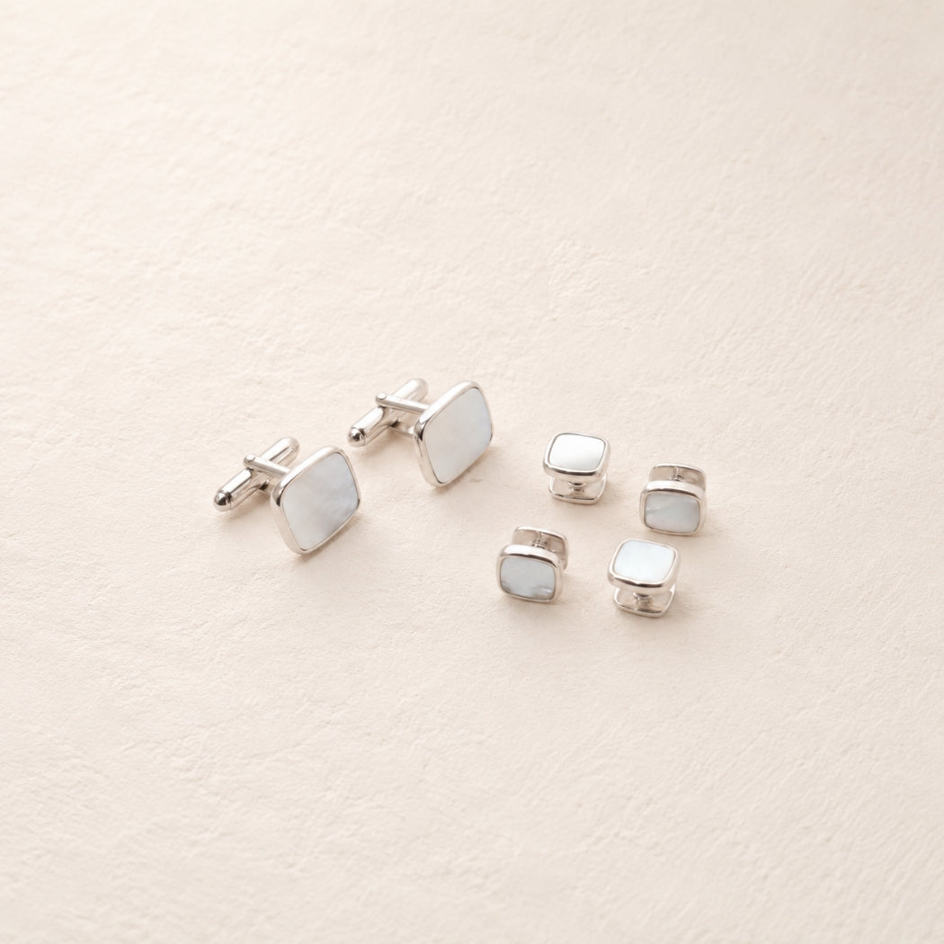 A set of silver-toned men's accessories including cufflinks and square shirt studs, each featuring a smooth, pale iridescent inlay, arranged on a textured cream background.
