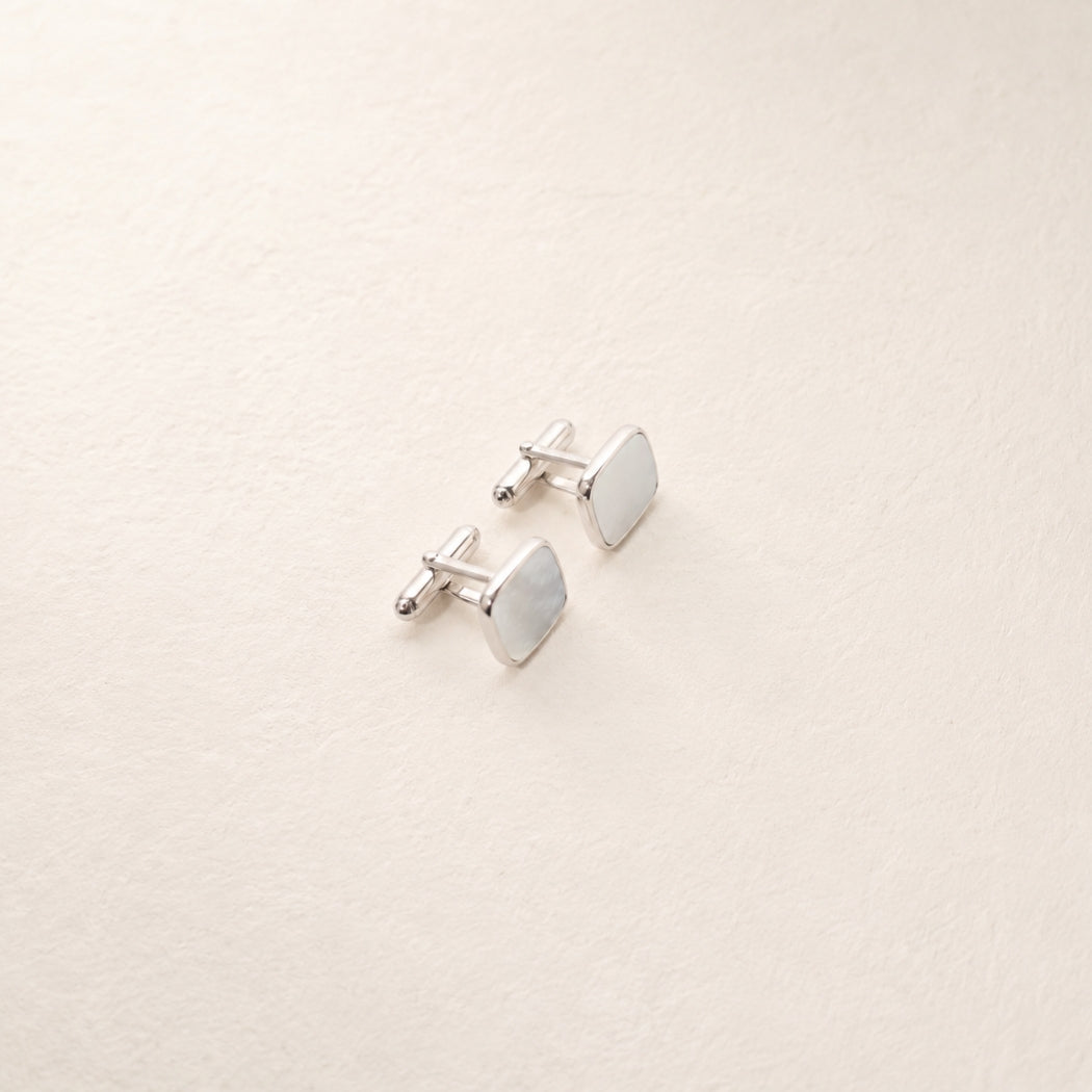 A pair of silver-toned square cufflinks with mother of pearl inlay, displayed on a textured off-white background.