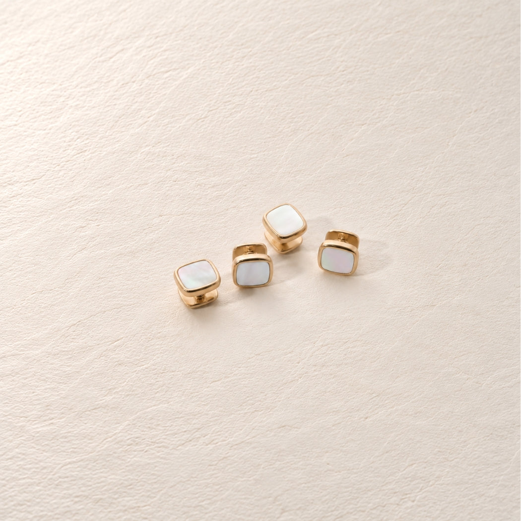 Four small square shirt stud with mother of pearl inlay and gold-tone edges, arranged on a light beige textured surface.