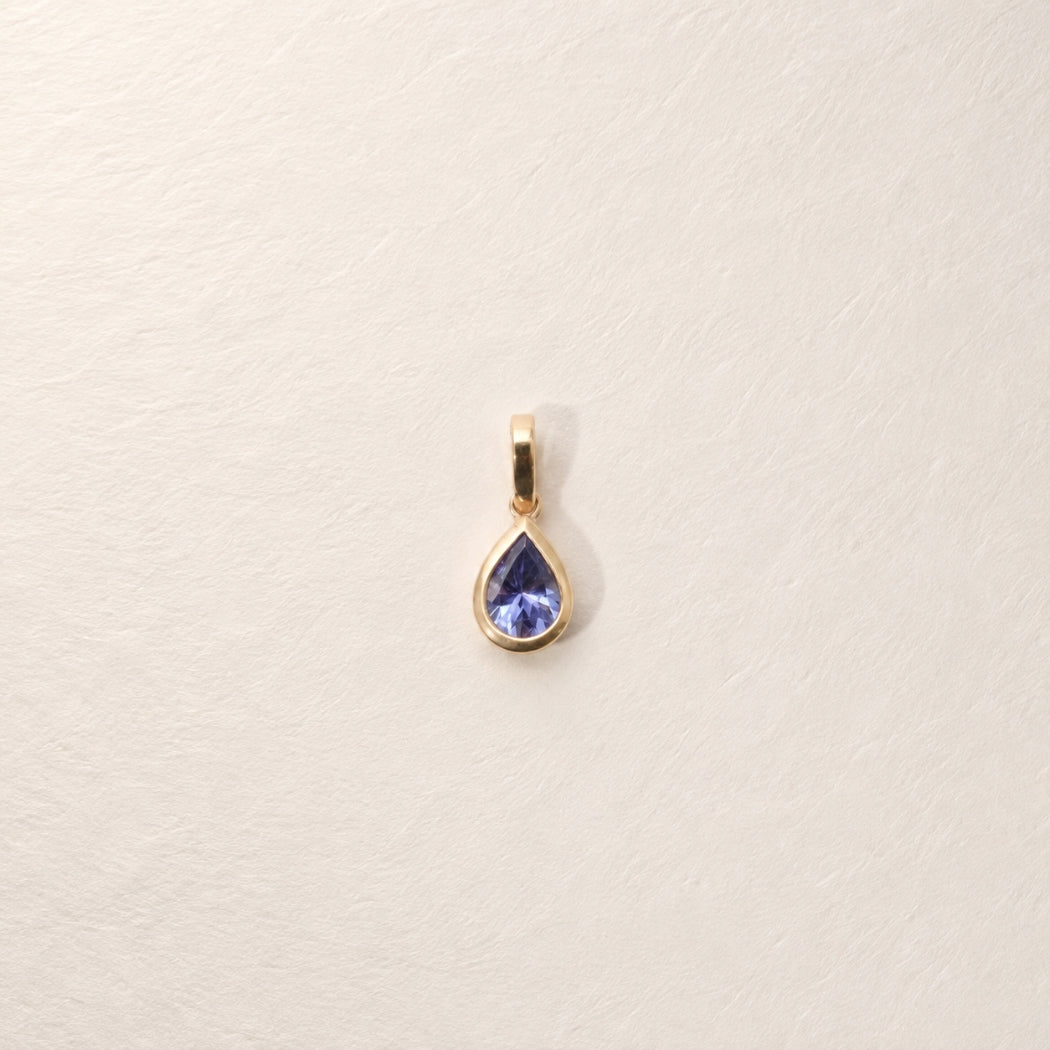 A gold teardrop pendant with a tanzanite blue gemstone, centered on a textured off-white background.
