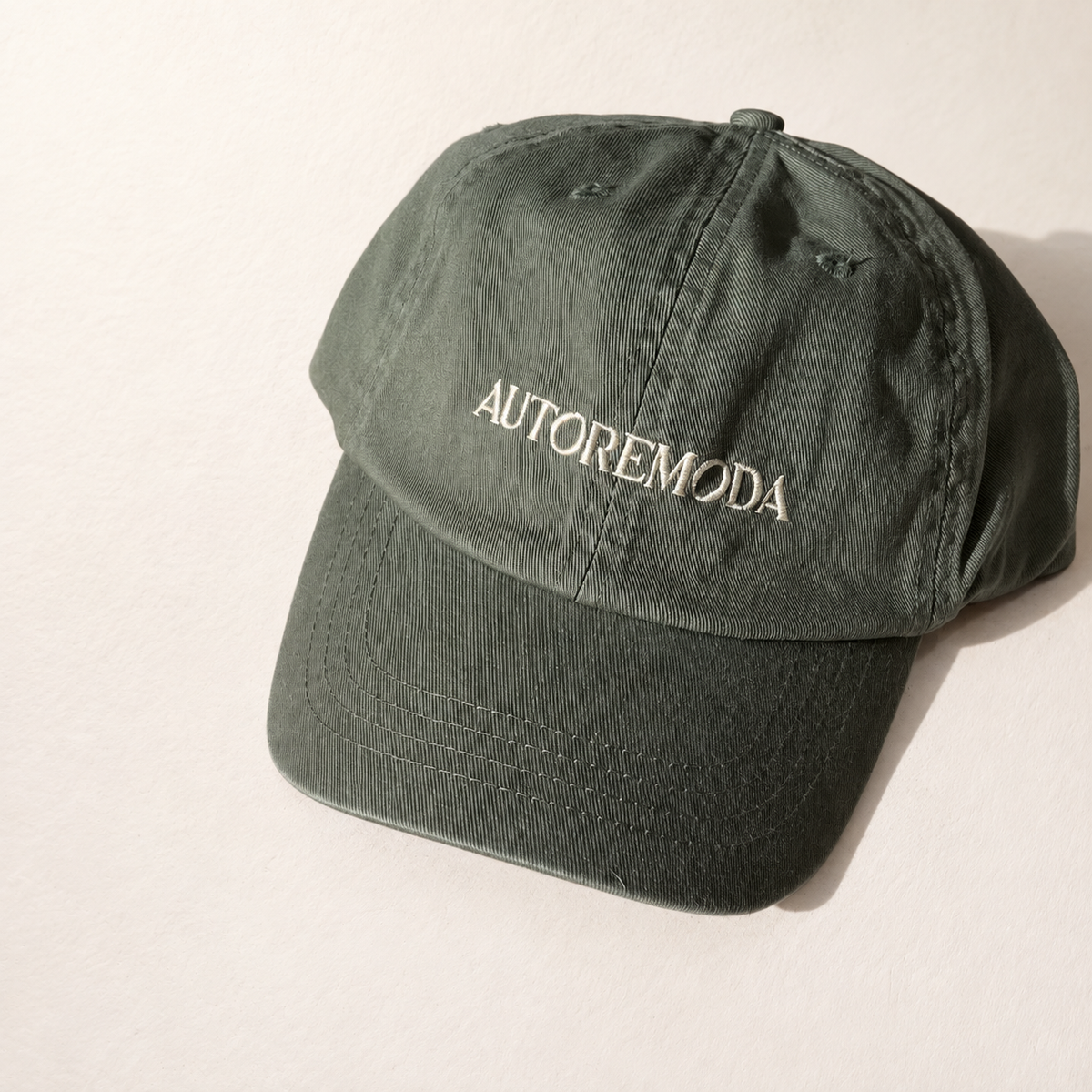 An olive green cotton baseball cap with 'AUTOREMODA' embroidered in white on the front, photographed on a light beige background.