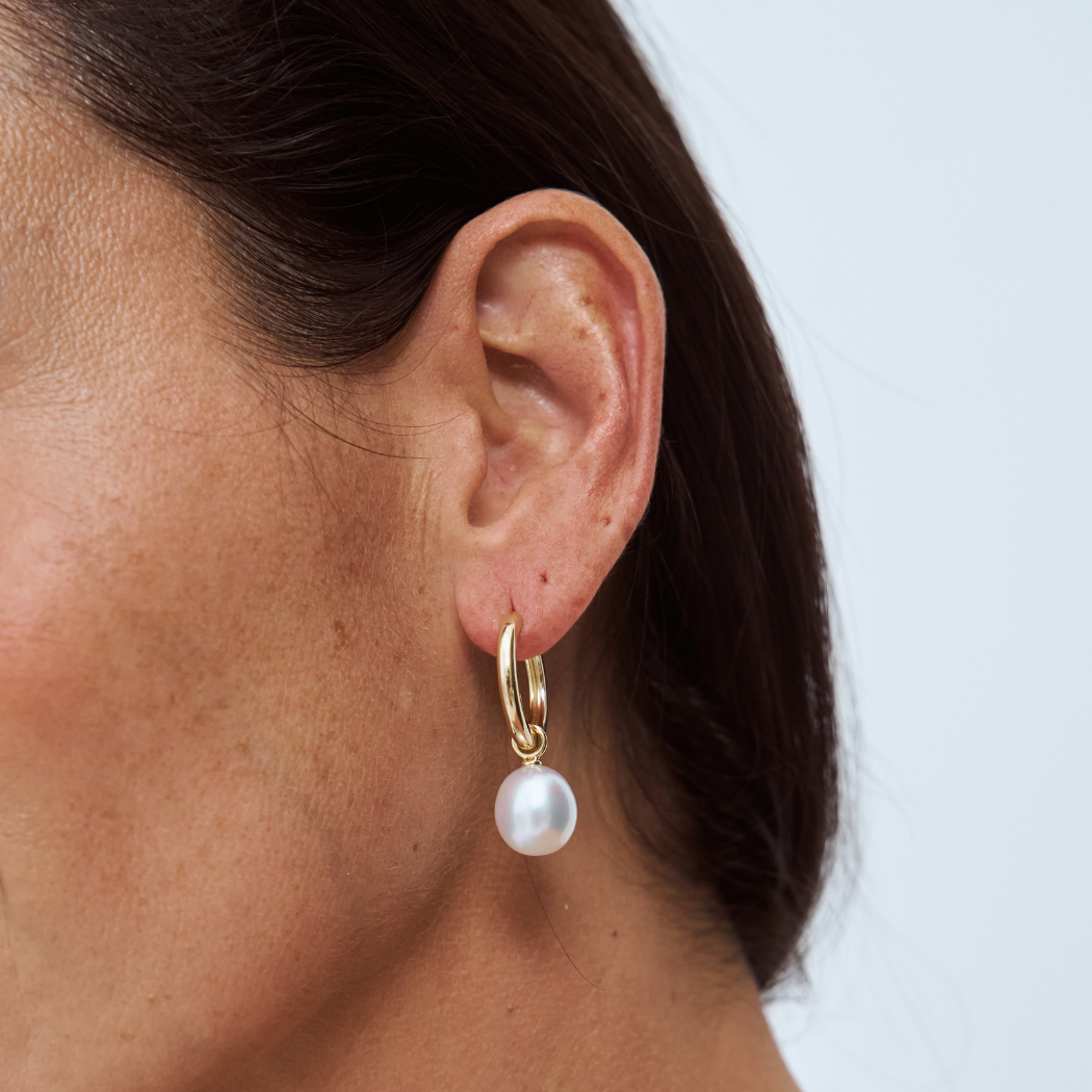 Close-up of an ear wearing a 9k yellow gold hoop earring with a south sea pearl pendant on a light background
