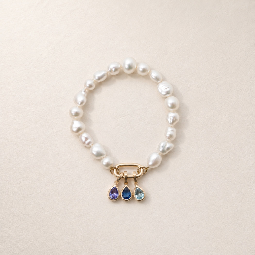 A south sea keshi pearl bracelet with a gold clasp and three dangling teardrop charms in aquamarine, tanzanite and sapphire displayed on a light beige textured background.
